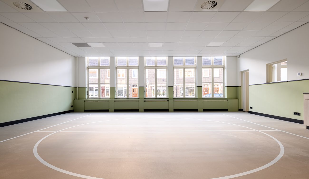 Lichte gym-/multizaal met mintgroene lambrisering, hoge ramen en cirkelbelijning op de vloer