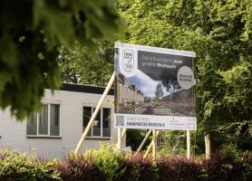 Bouwbord met impressie van nieuwbouwproject naast bestaand gebouw tussen groene bomen