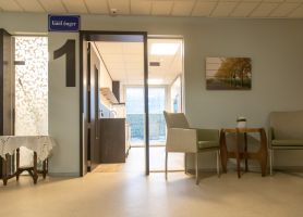 Zithoek in zorgcentrum met twee fauteuils en houten bijzettafel; links deur met groot cijfer 1 naar een lichte kitchenette, aan de muur een foto van een laan.