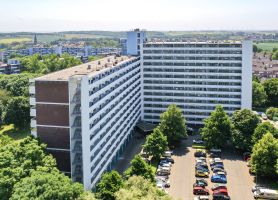 Luchtfoto van L-vormige galerijflat Veliahof in Maastricht met witte gerenoveerde gevels en lange balkonstroken, omringd door bomen en parkeerplaatsen.