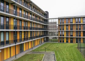 Uitzicht op het binnenterrein van een L-vormig appartementencomplex; ritmische gele en oranje gevelstroken, galerijen met zwarte hekwerken en een verzorgd grasveld.