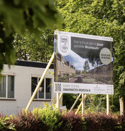 Bouwbord met impressie van nieuwbouwproject naast bestaand gebouw tussen groene bomen