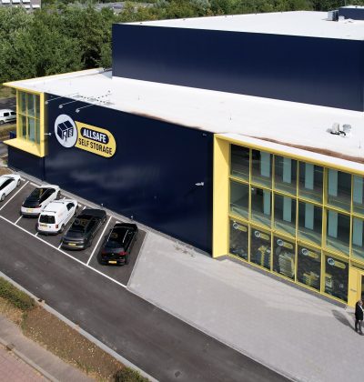 Luchtfoto van de nieuwbouw van Allsafe Self Storage in Maastricht: donkerblauw volume met felgele kaders rond de glazen hoekgevel, witte dakrand en parkeervakken met auto’s langs de voorzijde; twee personen lopen richting de entree, omringd door wegen en groen.