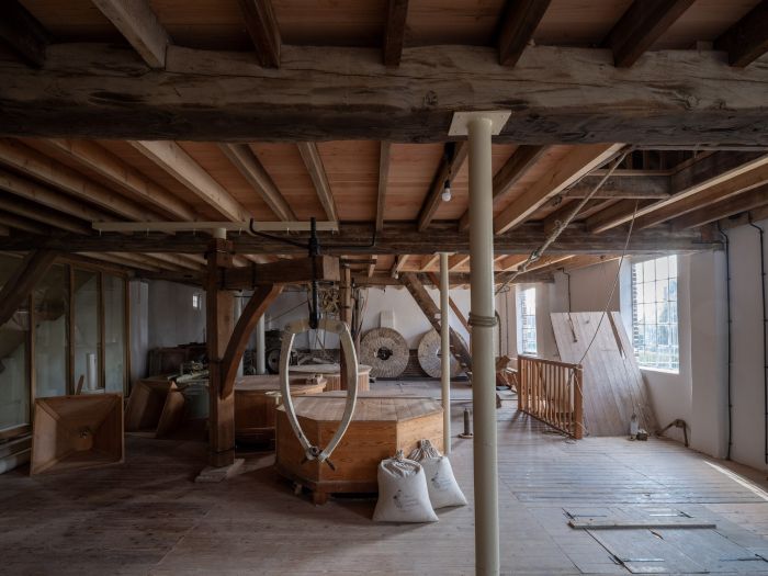 Interieur Grathemermolen – maalzolder met houten balken, maalinstallatie en -stenen, graanzakken en daglicht via roosterraam.