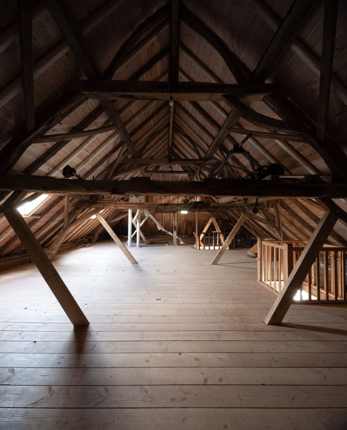Zolder van de Grathemermolen met open houten spantconstructie onder het zadeldak, brede houten vloer en zicht op delen van het maalwerk; trapleuning rechts.