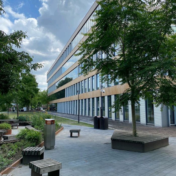 Modern kantoorgebouw met langgerekte glazen gevel en houten accenten, omgeven door bomen, zitbanken en een betegeld plein onder een deels bewolkte lucht.