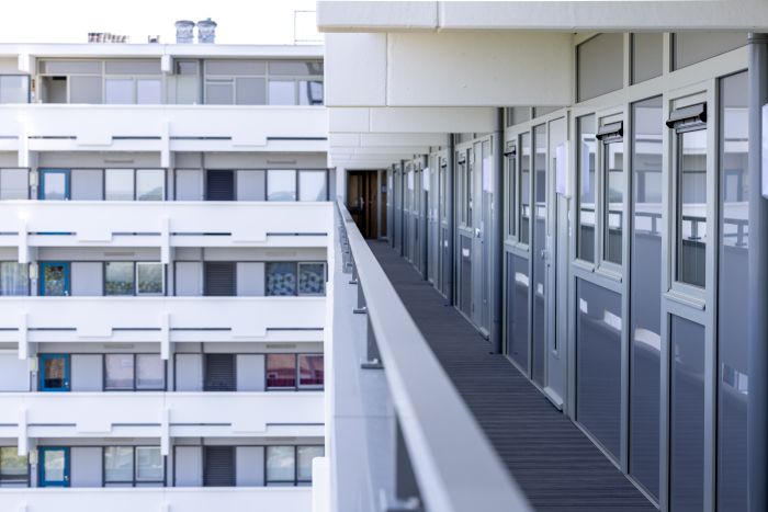 Perspectief langs een galerij met grijze voordeuren en glazen balustrade; aan de overzijde is de witte gevel met ramen en balkons van de tegenoverliggende vleugel te zien
