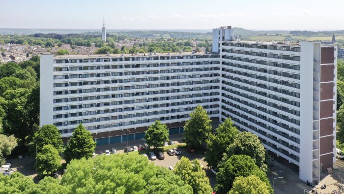 Luchtfoto van L-vormige galerijflat Veliahof in Maastricht, met witte gerenoveerde gevels en lange balkonstroken, omgeven door bomen en parkeerplaatsen.