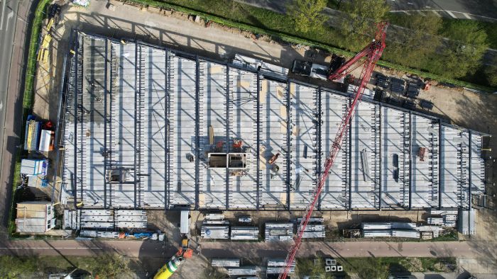 Verticale dronefoto van een bouwplaats met een trapeziumvormig gebouw in aanbouw: raster van stalen/ prefab betonnen liggers, rood kraangiek diagonaal over het beeld, stapels prefab elementen langs de randen en enkele bouwvakkers zichtbaar op de vloer.