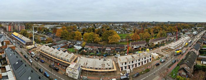 Luchtfoto panorama van nieuwbouw rijwoningen – bouwblokken met steigers en kranen langs bochtige straat in woonwijk, herfstbomen