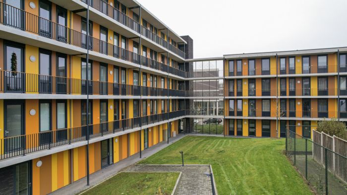 Uitzicht op het binnenterrein van een L-vormig appartementencomplex; ritmische gele en oranje gevelstroken, galerijen met zwarte hekwerken en een verzorgd grasveld.