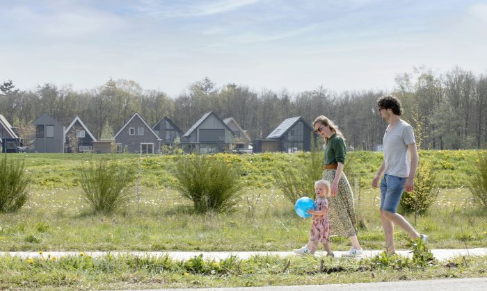 Gezin wandelt langs groene strook met peuter en blauwe bal, moderne woningen met zadeldaken en bosrand op achtergrond