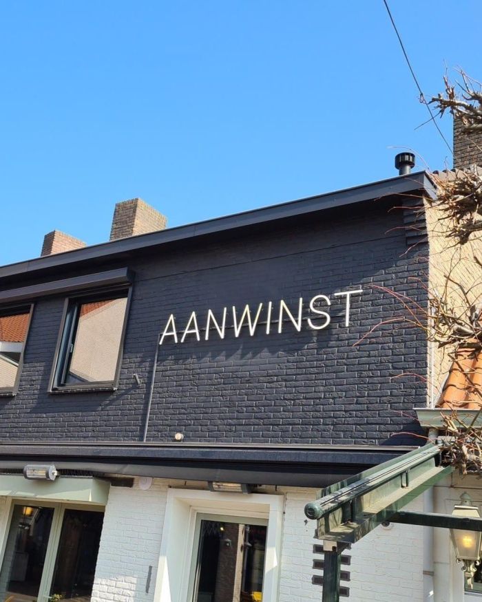 Gevel van eetcafé De Aanwinst met zwart geschilderde baksteen en grote verlichte letters “AANWINST”, onder een helderblauwe lucht.
