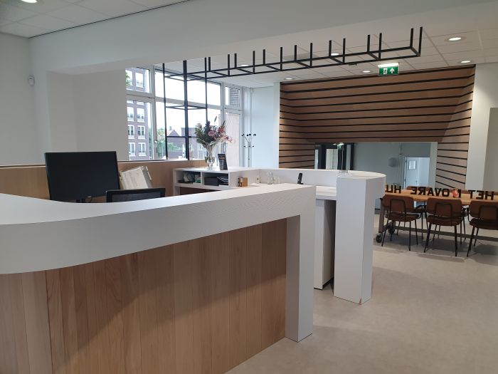 Ontvangstbalie met gebogen wit-houten front in een lichte kantoorruimte; groot raam met daglicht, zwart plafondrooster, houten lattenwand met doorgang en daarachter een vergadertafel met stoelen en glazen deur.