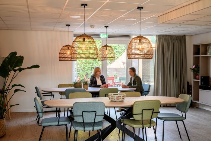 Ontmoetingsruimte met ovale tafels, rieten hanglampen, twee vrouwen in overleg, zicht op binnentuin.