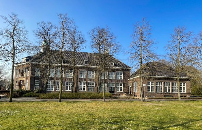 Verbouwing Huize Padua in Boekel – monumentaal gebouw met klassieke bakstenen gevels en hoge ramen, gezien vanaf het park met bomen en grasveld onder een heldere blauwe lucht.