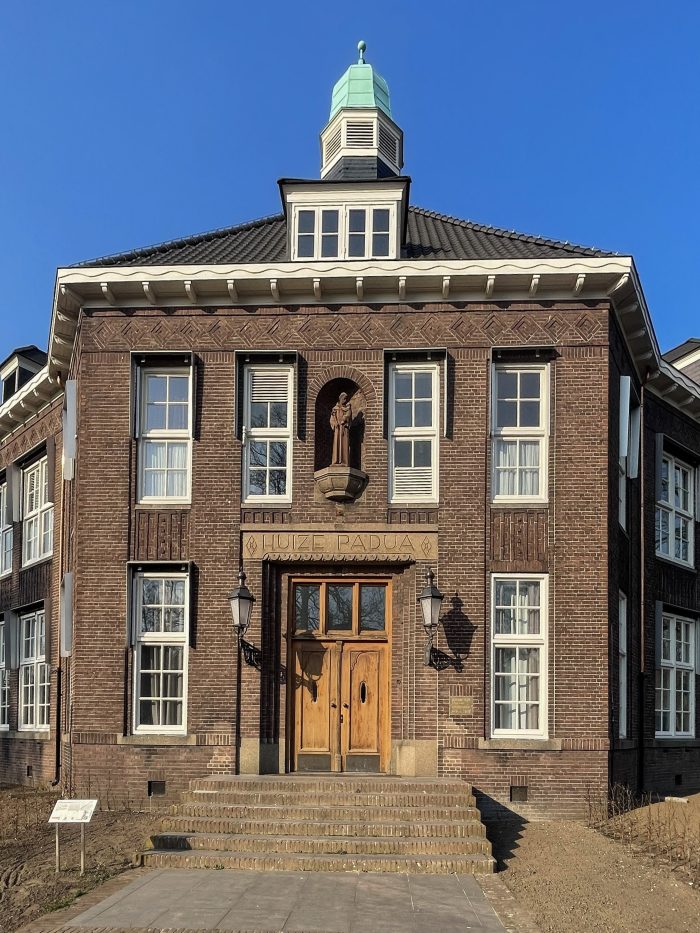 Verbouwing Huize Padua Boekel – close-up van de monumentale hoofdentree met houten dubbele deur, beeld in nis boven het opschrift “HUIZE PADUA”, bakstenen gevel en trapportaal onder een helderblauwe lucht.