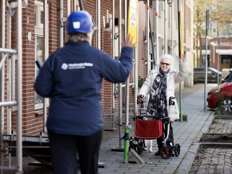Een bewoonster zwaait vrolijk naar een medewerker van het bouwbedrijf. De medewerker met bouwhelm zwaait terug