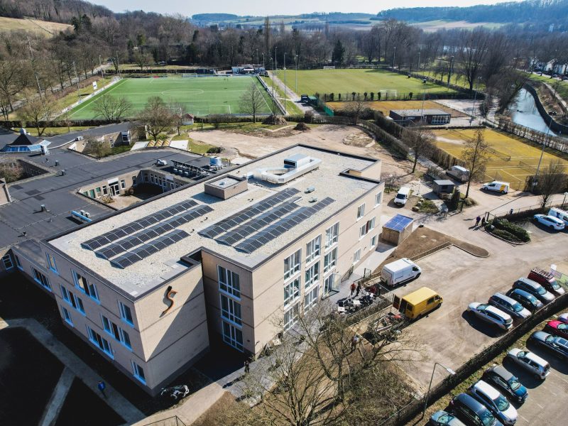 Luchtfoto van een modern gebouw met zonnepanelen op het dak, parkeerplaats en sportvelden ernaast.