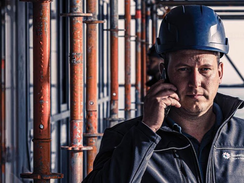 Man met blauwe bouwhelm belt op een bouwplaats terwijl hij tussen steigerelementen staat.